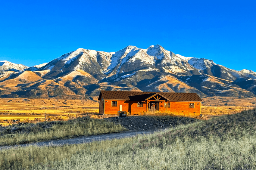 one of Montana vacation rentals, a cabin, in the foreground, with snow-capped mountains in the background, all set against a bright blue sky.